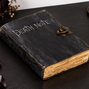 Op de afbeelding: Een zwart rechthoekig notitieboek met de woorden DEATH NOTE in het wit op de voorkant gedrukt. Het boek heeft een verweerde textuur, een messingkleurige metalen sluiting aan de zijkant en dikke, verouderd ogende pagina's met ruwe randen.