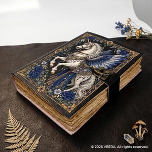 Op de afbeelding: Een dagboek met een zwarte harde kaft, voorzien van een witte gevleugelde eenhoorn, blauwe rozen en gouden filigraanaccenten. Het boek heeft een metalen sluiting en verouderde papieren pagina's met ruwe randen. De tekst 2026 VEERA. All rights reserved. is zichtbaar.