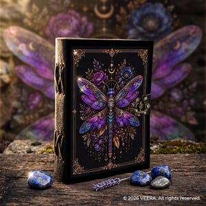 May include: A black journal featuring a vibrant purple and blue dragonfly illustration with gold accents, a metal latch closure, and side leather lacing. The cover includes the text 2026 VEERA. All rights reserved. at the bottom right corner.