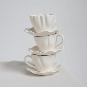 Może przedstawiać: Pionowy stos trzech matowych białych filiżanek ceramicznych z pasującymi spodkami. Każda filiżanka ma wyraźną, żłobkowaną teksturę przypominającą płatki kwiatów na zewnętrznych ściankach oraz spodek z falistym, wyciętym brzegiem.