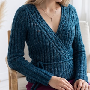 Peut inclure: Un pull portefeuille bleu sarcelle profond avec un motif en tricot côtelé texturé et des taches multicolores subtiles. Le vêtement présente des manches longues, une encolure en V et un lien à nouer à la taille, accompagné d'un collier en argent.