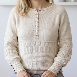 May include: A cream-colored long-sleeve knit sweater featuring a round neckline with a vertical placket of six silver-toned buttons. The garment includes textured diamond-patterned panels across the chest and sleeves, with three matching buttons on each cuff.