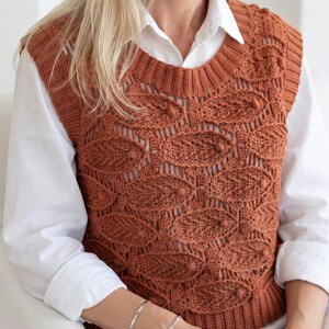 May include: A rust-coloured sleeveless sweater vest featuring a repeating crochet leaf pattern with a ribbed crew neckline and hem. The garment is worn over a crisp white long-sleeved button-down shirt, showcasing the intricate textured stitch work.