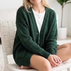 Könnte beinhalten: Eine dunkelwaldgrüne Strickjacke mit V-Ausschnitt, langen Ärmeln mit gerippten Bündchen, einer durchgehenden Knopfleiste mit farblich passenden Knöpfen und zwei aufgesetzten Vordertaschen. Die Strickjacke wird über einem schlichten weißen T-Shirt getragen.