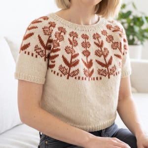 May include: A short-sleeved knit sweater in cream with a round neckline and ribbed trim. The yoke features a contrasting rust-colored floral pattern with vertical stems and circular flower heads arranged in a repeating design around the chest.