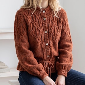 May include: A rust-colored knit cardigan featuring a textured cable knit pattern on the upper front and sleeves. The garment has a round neckline, long sleeves with ribbed cuffs, a button-down front with light-colored buttons, and a drawstring waist.