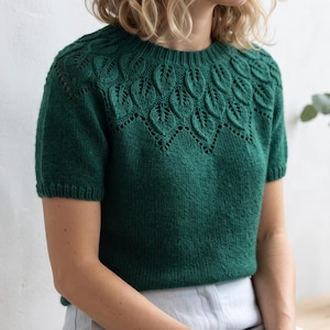 May include: A short-sleeved, dark forest green knit sweater featuring a round neckline and a decorative yoke pattern composed of intricate, raised leaf-shaped motifs with delicate eyelet detailing along the lower edge of the design.