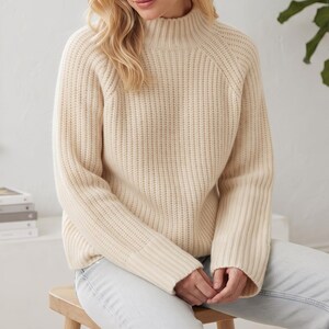 May include: A cream-colored long-sleeve sweater featuring a ribbed mock neck, vertical ribbed knit texture throughout the body, and wide ribbed cuffs at the sleeves. The garment has raglan sleeves and a straight hemline, providing a structured fit.