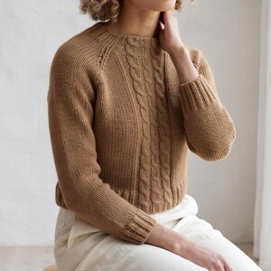May include: A tan long-sleeve sweater featuring a round neckline, ribbed cuffs, and a ribbed hem. The front of the garment displays a vertical cable knit pattern running down the center, contrasted by smooth stockinette stitch sleeves and sides.