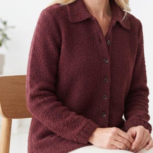 May include: A long-sleeved, button-down shirt in a deep burgundy color. The garment features a textured, bouclé-like fabric, a classic collar, and matching dark buttons running down the front placket. The cuffs are finished with a ribbed knit trim.