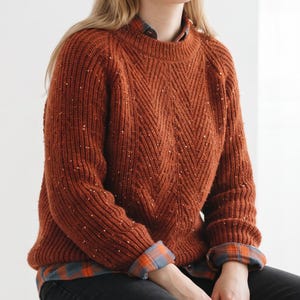 Könnte beinhalten: Ein rostfarbener, grob gestrickter Pullover mit vertikalem Zickzackmuster und feinen Sprenkeln in helleren Farben. Der Pullover hat einen Rundhalsausschnitt und lange Ärmel und wird über einem karierten Hemd in Orange- und Blautönen getragen.