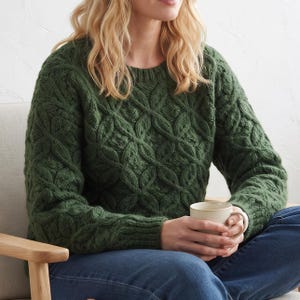 Könnte beinhalten: Ein waldgrüner Langarmpullover mit einem strukturierten, erhabenen Gitter-Zopfmuster auf der Vorderseite und den Ärmeln, einem Rundhalsausschnitt und gerippten Bündchen. Das Kleidungsstück wird mit einer dunkelblauen Jeans und einer Keramiktasse kombiniert.