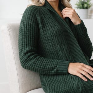 May include: A dark forest green open-front cardigan featuring a waffle knit texture on the sleeves and back, complemented by a vertical cable knit pattern along the shawl collar and front edges, with long sleeves and ribbed cuffs.