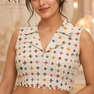 Puede incluir: Una blusa sin mangas de color blanco roto con cuello y un patrón de cuadrícula bordado con pequeñas flores multicolores en rojo, azul, verde y amarillo. El dobladillo está rematado con una hilera de pequeños pompones blancos.