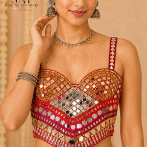 May include: A sleeveless red blouse featuring a sweetheart neckline and intricate mirror work embroidery. The garment is adorned with small reflective circular mirrors arranged in a geometric pattern, accented by vibrant red and orange thread stitching.
