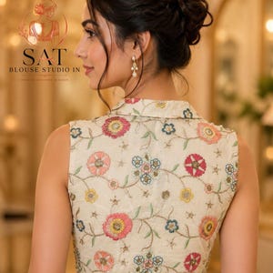 Puede incluir: Una blusa sin mangas de color crema con cuello y un patrón de bordado floral en tonos rosa, amarillo y azul. El dobladillo está rematado con una hilera de pequeñas cuentas blancas. El texto SAT BLOUSE STUDIO IN es visible.