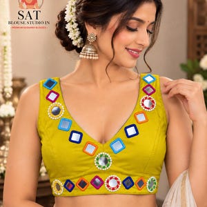 May include: A sleeveless chartreuse blouse featuring a deep V-neckline adorned with square and circular mirrors framed by blue, red, and green borders. The garment includes a horizontal band at the waist decorated with additional mirrors and beads.