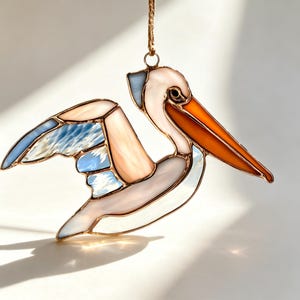 Op de afbeelding: Een glas-in-lood pelikaanornament met een lange oranje snavel, een lichtroze kop en lichaam, en vleugels met doorschijnende blauwe en witte glazen panelen. De onderdelen zijn verbonden door dunne koperkleurige metalen soldeerlijnen.