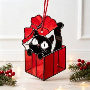 Puede incluir: Un adorno colgante de vidrieras que muestra un gato negro con grandes ojos blancos asomándose desde una caja de regalo roja. El gato tiene un gran lazo rojo encima y está suspendido por un cordel rojo. Tiene contornos de metal negro.