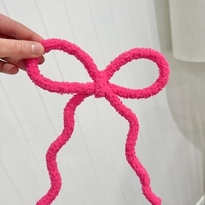 May include: A bright hot pink bow crafted from a fuzzy, textured yarn material. The bow features two symmetrical loops at the top and two long, slightly wavy tails hanging downwards, creating a soft and tactile appearance in a vibrant shade.