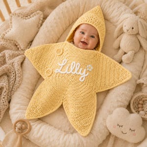 Peut inclure: Une couverture d'emmaillotage pour bébé en forme d'étoile jaune, réalisée en fil crocheté. Le centre de la couverture présente le prénom Lilly brodé en écriture cursive blanche, accompagné d'une petite fleur blanche et de deux boutons ronds.