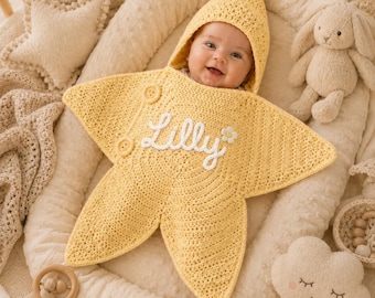 Peut inclure: Une couverture d'emmaillotage pour bébé en forme d'étoile jaune, réalisée en fil crocheté. Le centre de la couverture présente le prénom Lilly brodé en écriture cursive blanche, accompagné d'une petite fleur blanche et de deux boutons ronds.