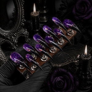 May include: A set of ten stiletto-shaped press-on nails featuring a deep purple and black gradient base with embedded iridescent glitter flakes. The nails are arranged in two rows on a wooden display stand, showcasing a sharp, pointed tip design.
