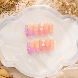 Könnte beinhalten: Ein Set aus zehn künstlichen Fingernägeln mit einem Farbverlauf von zartem Pfirsich am Nagelbett bis zu kräftigem Pink an der Spitze. Die Nägel sind mit dreidimensionalen lavendelfarbenen Blütenblättern und kleinen klaren Strasssteinen verziert.