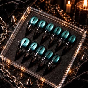 May include: A set of ten stiletto-shaped press-on nails arranged in a clear plastic display case. Each nail features a deep teal, cat-eye magnetic shimmer at the base that transitions into a sharp, translucent black pointed tip with a glossy finish.