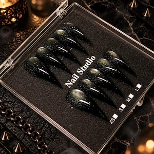 May include: A set of ten stiletto-shaped press-on nails in a clear plastic storage case. The nails feature a dark black base with a shimmering green cat-eye magnetic effect. The case is labeled with the text Nail Studio and size indicators.