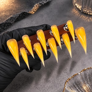 May include: A set of ten stiletto-shaped press-on nails featuring a translucent, vibrant yellow finish. The nails are arranged in two rows on a dark wooden display bar, showcasing a sharp, pointed tip and a smooth, glossy surface texture.