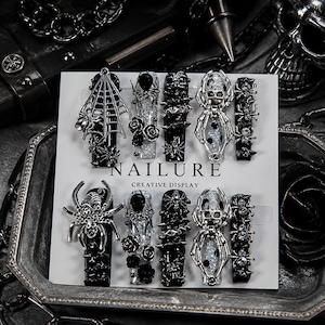 May include: A set of ten press-on nails on a white display card labeled NAILURE CREATIVE DISPLAY. The nails feature black and silver glitter finishes, adorned with three-dimensional silver spider, spiderweb, rose, and skull embellishments.