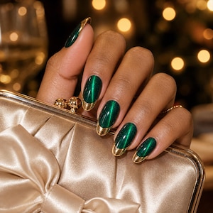 Könnte beinhalten: Eine champagnerfarbene Satin-Clutch mit Schleifendetails, gehalten von Fingern mit mandelförmigen Nägeln, die in einem tiefen Smaragdgrün mit Katzenaugen-Effekt lackiert sind und an jeder Nagelspitze einen reflektierenden Goldakzent aufweisen.