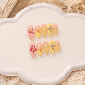 Könnte beinhalten: Ein Set aus zehn mandelförmigen Press-on-Nägeln mit einem Farbverlauf in Zartrosa und Gelb. Die Designs umfassen handgemalte rote Blumenmotive, goldene sternförmige Anhänger, kleine goldene Perlen und silberfarbene Kreuzanhänger mit Kristallen.