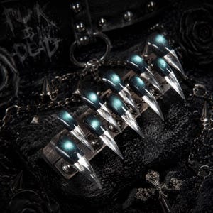 May include: A set of ten press-on nails featuring a stiletto shape with a dark, metallic teal finish at the base that transitions into a clear, transparent tip. The nails are arranged in a row on a rectangular display stand with silver studs.