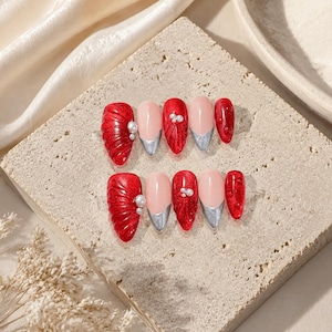 May include: A set of ten press-on nails featuring a mix of designs including solid red with textured shell-like ridges, nude bases with metallic silver tips, and red nails accented with small white pearls and raised circular dots in a glossy finish.