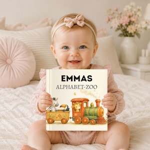 Könnte beinhalten: Ein Kinderbuch mit festem Einband mit dem Titel EMMAS ALPHABET-ZOO, das eine farbenfrohe Illustration eines Dampfzuges zeigt, der ein Zebra, einen Tukan und eine Giraffe befördert. Das Buchcover hat einen cremefarbenen Hintergrund mit schwarzem Text.