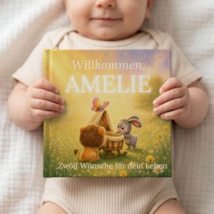 Könnte beinhalten: Ein quadratisches Hardcover-Buch mit einem hellen Einband, der einen Löwen, einen Hasen und einen Schmetterling an einer Wiege auf einer Wiese zeigt. Der Text Willkommen, AMELIE und Zwölf Wünsche für dein Leben steht in Weiß auf gelbem Grund.
