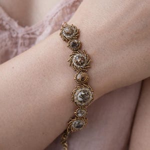 Puede incluir: Una pulsera que presenta una serie de piedras redondas jaspeadas en tonos marrón y blanco, cada una rodeada por un borde decorativo de pequeñas cuentas metálicas de color bronce. Las piedras varían en tamaño y están unidas en línea.