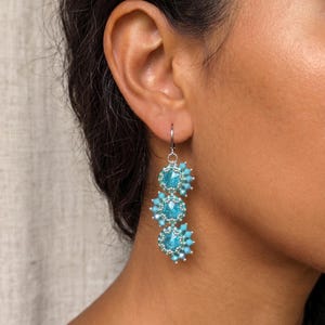Può includere: Un orecchino pendente caratterizzato da tre componenti circolari allineati verticalmente in un colore blu turchese brillante. Ogni componente è centrato con un cristallo sfaccettato circondato da un bordo di piccole perline, sospeso a un gancio in metallo argentato.