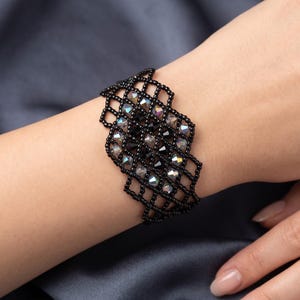 Könnte beinhalten: Ein breites Armband mit einem schwarzen Perlen-Gittermuster. Das Design enthält facettierte, schillernde Kristalle, die in die Mitte des Gitters eingearbeitet sind und ein strukturiertes, geometrisches Erscheinungsbild erzeugen.