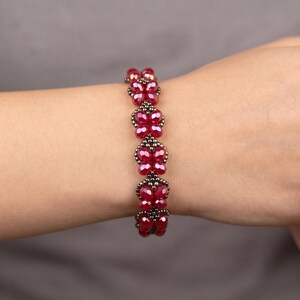 May include: A bracelet composed of a series of deep red, faceted crystal butterfly-shaped beads linked together with small, dark metallic seed beads. The design features a repeating pattern of these red butterfly motifs connected by dark beads.