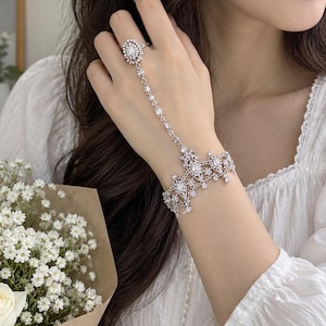 May include: A silver-toned hand chain featuring a delicate bracelet connected to a ring by a slender chain. The piece is adorned with numerous clear, sparkling crystals set in an intricate, filigree-style metal design that covers the wrist and finger.