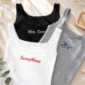 Puede incluir: Tres camisetas de tirantes acanaladas con ribete de encaje y pequeños lazos en el escote. La camiseta blanca lleva el texto rojo Seraphina, la negra muestra el texto blanco Mrs. Emma y la gris presenta el texto bordado azul oscuro ZOE.