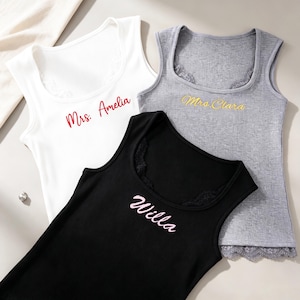 Può includere: Tre canotte senza maniche in bianco, grigio e nero, ciascuna con bordo in pizzo sullo scollo e sull'orlo. Il top bianco mostra il testo ricamato in rosso Mrs. Amelia, quello grigio il testo giallo Mrs. Clara e quello nero il testo rosa Willa.