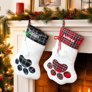 May include: Two white plush paw-shaped Christmas stockings with tartan cuffs. The left stocking has a green tartan cuff with the name Daniel and a green ribbon. The right stocking has a red tartan cuff with the name Audrey and a red ribbon.
