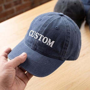 May include: A navy blue baseball cap with a washed-out finish featuring the word CUSTOM embroidered in white capital letters on the front panel. The cap has a curved brim, a button on the top, and stitched eyelets for ventilation.