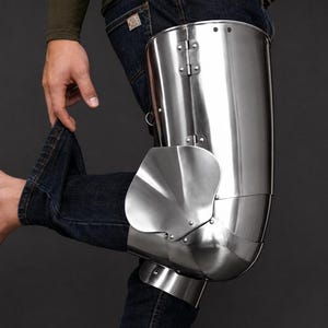 May include: A polished steel leg armor piece covering the thigh and knee, featuring a hinged construction with a rounded knee cap and a side wing plate. The metal surface is smooth and reflective, secured with visible rivets and metal hinges.