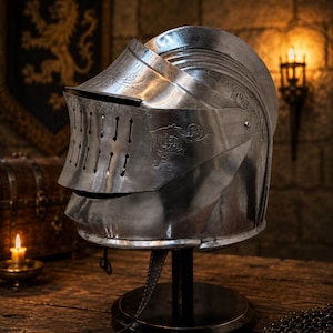 Peut inclure: Un casque de chevalier en acier poli doté d'une visière articulée avec des fentes de ventilation verticales et des gravures florales complexes sur le côté. Le casque est monté sur un support circulaire noir avec une jugulaire en cuir.