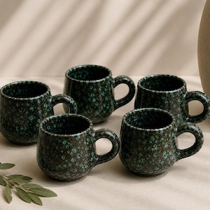 Op de afbeelding: Een set van vijf kleine keramische mokken met een donkerbruine basis en een gespikkeld glazuur in groen en blauwgroen. Elke mok heeft een afgerond lichaam en een dik, gebogen handvat. De mokken zijn in een groep bij elkaar geplaatst.